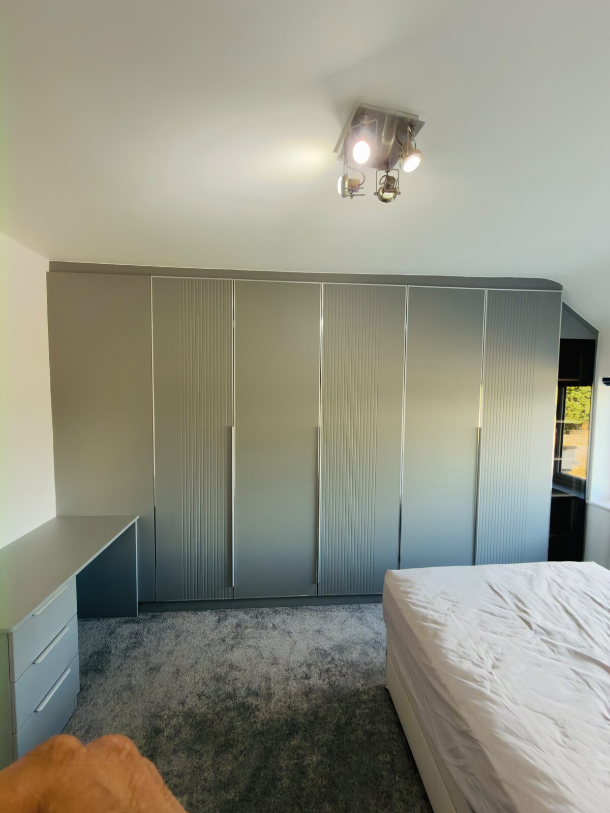 Six-door fitted wardrobe run in greige, mixing flat and fluted door panels, with a curved radiused corner section following a sloping ceiling, and a matching desk and drawer unit