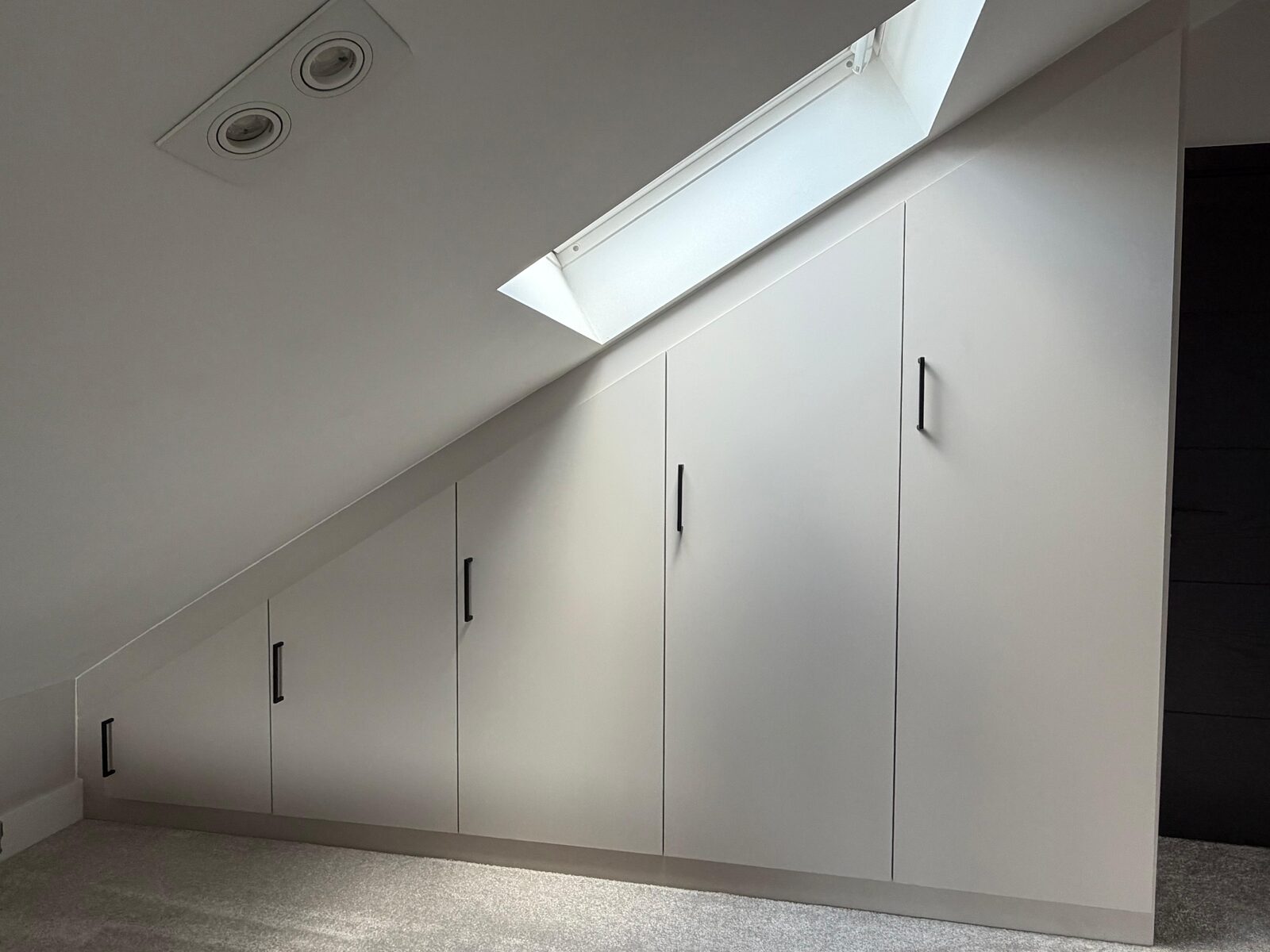 Five fitted wardrobe doors of graduating height following the slope of a loft ceiling, with black bar handles and a Velux window above