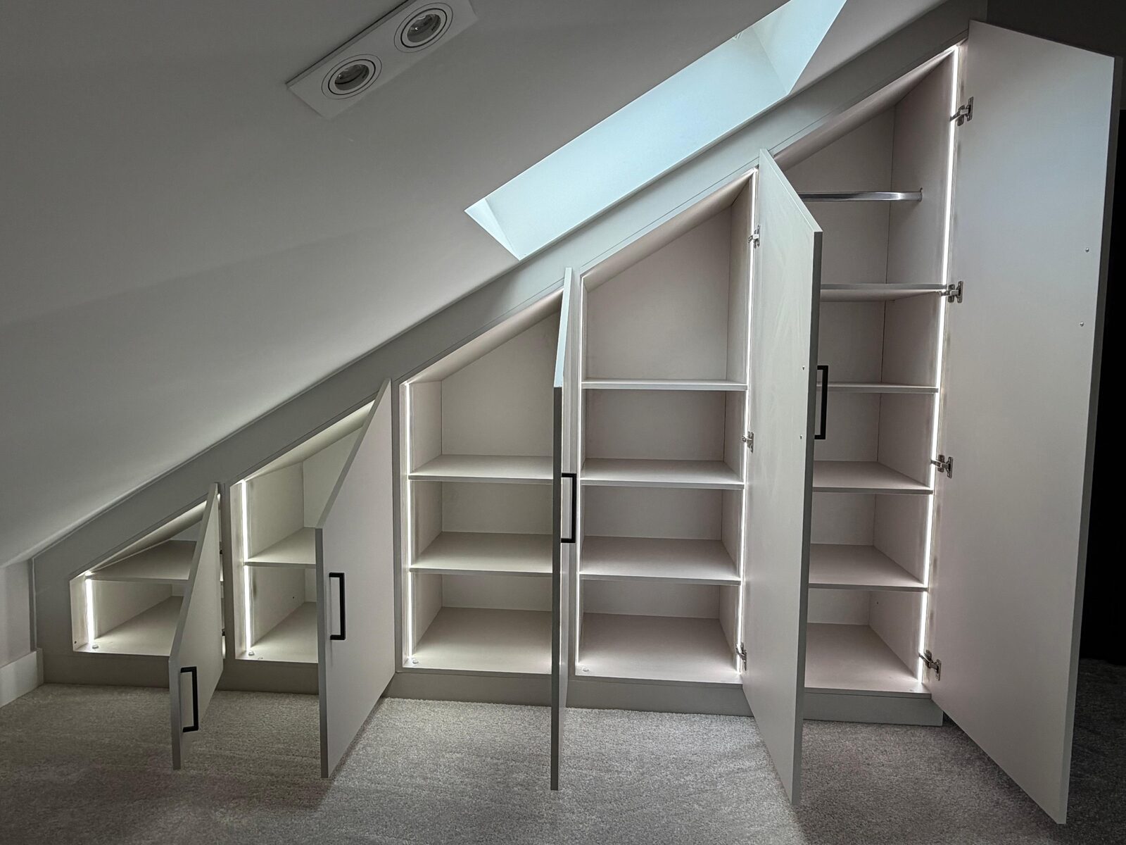 Loft eaves wardrobe with doors open, showing graduated shelves cut to the roof pitch, a hanging rail at the full-height end and LED strip lighting in each bay