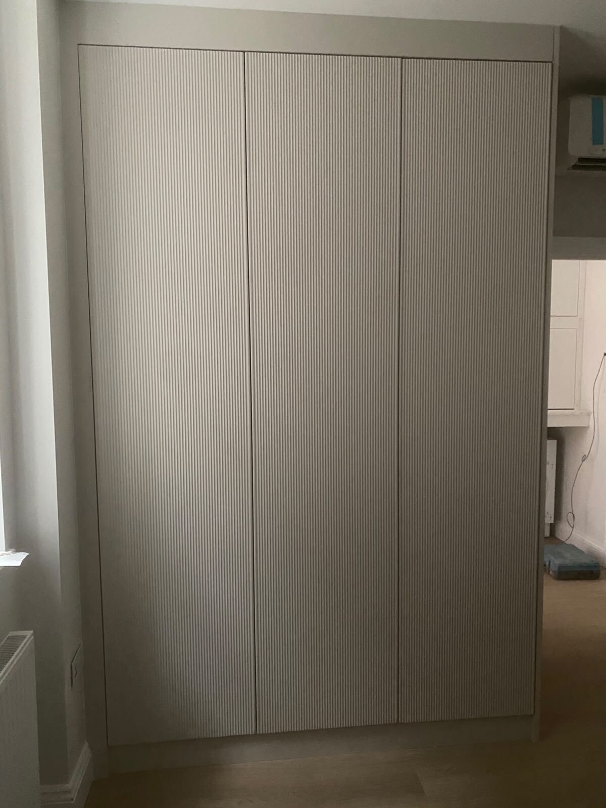 Three-door fluted fitted wardrobe in pale greige by UKB, running floor to ceiling with handleless doors