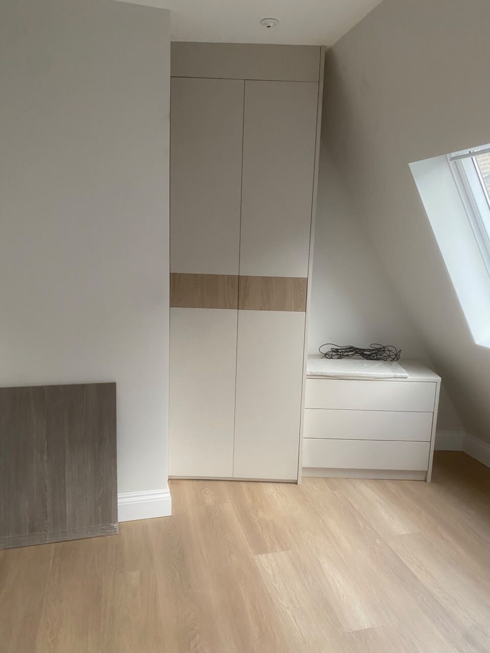 Cream fitted wardrobe with a horizontal oak-effect band across the doors, beside a chest of drawers under a sloping ceiling