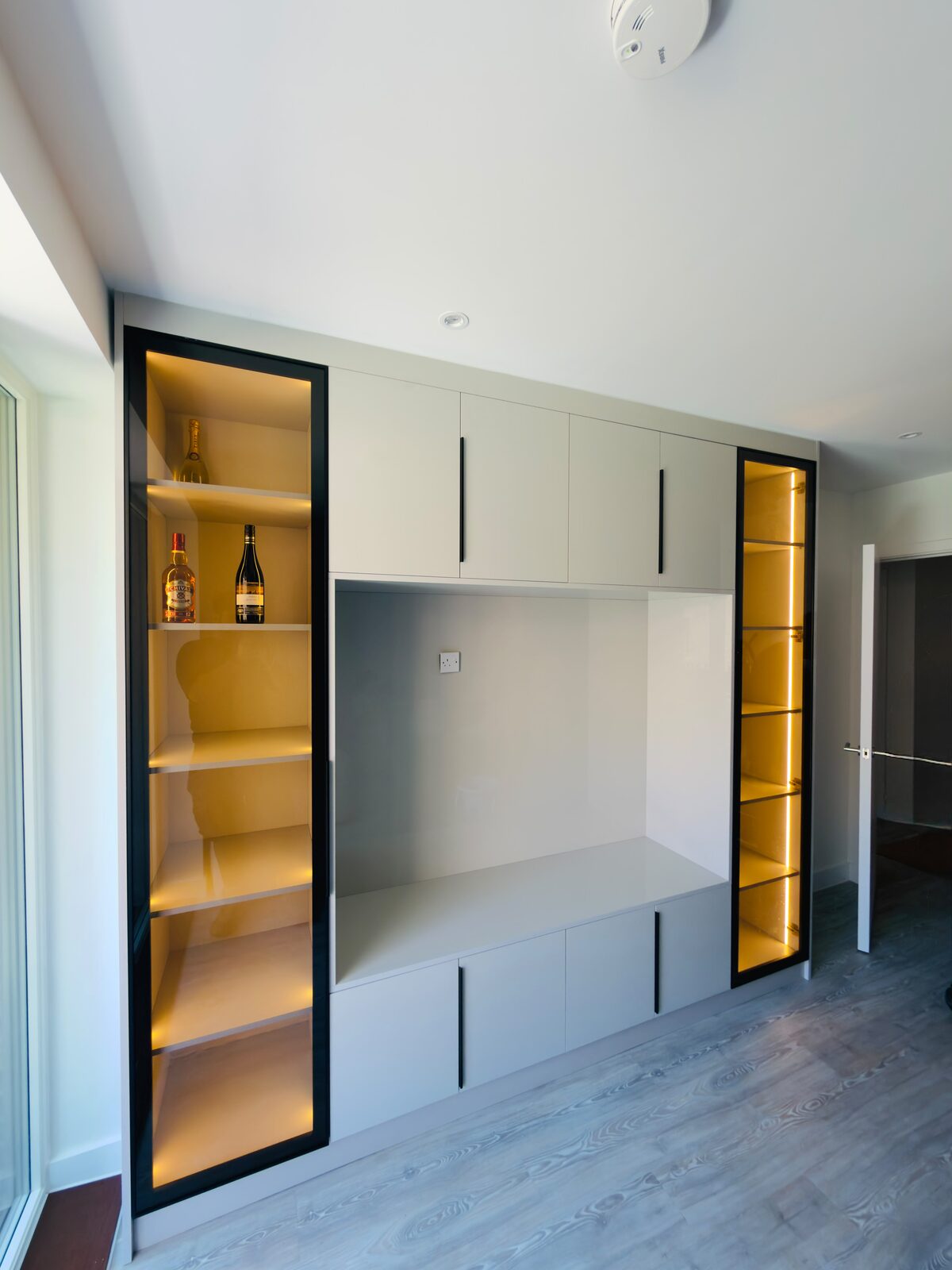 Living room media wall with lit black-framed glass display cabinets either side of a television recess and drawers below